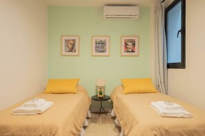 Una habitación con dos camas, cada una con una almohada amarilla y una toalla blanca, una mesita con una lámpara, y tres cuadros en la pared.