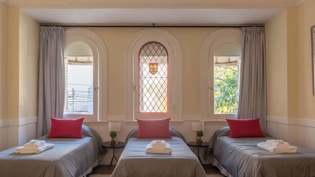 A hotel room with two beds, each with a grey bedspread and a red pillow, and a window with a decorative glass panel.