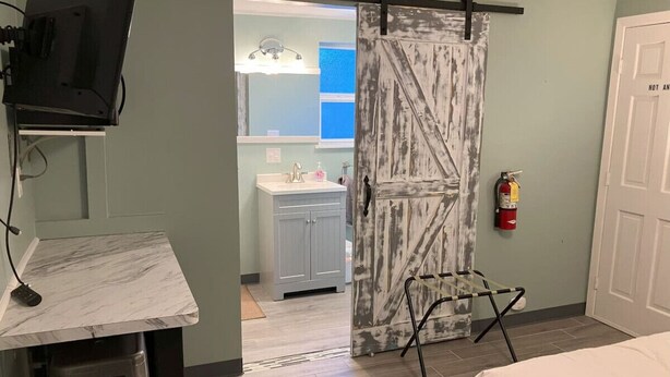 Ein Zimmer mit einem Bett, einem Schreibtisch mit Fernseher, einem Badezimmer mit Waschbecken und Spiegel sowie einem Feuerlöscher.
