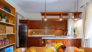 A kitchen with wooden cabinets, a stainless steel refrigerator, and a dining table set with a pitcher of orange juice, a bowl of fruit, and two glasses of orange juice. - Casa Loula (Corfu)