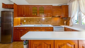 A kitchen with wooden cabinets, a stainless steel refrigerator, a white countertop, and a window with curtains. - Casa Loula (Corfu)