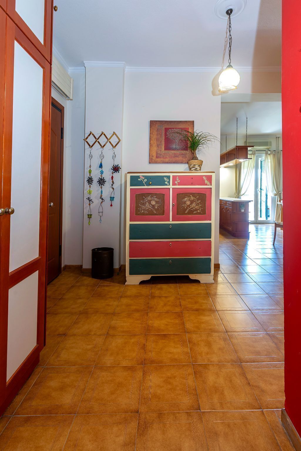A hallway with a wooden door, a colorful dresser, a hanging plant, and a painting on the wall.