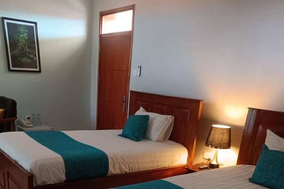 A hotel room with two wooden beds, each with a teal bedspread and white linens. There is a wooden nightstand with a lamp and a phone on the left side. A door is visible in the background.