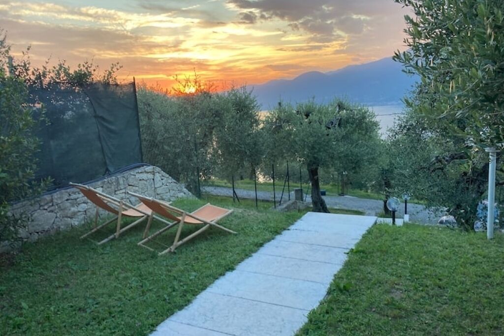 A garden with a stone wall, two wooden chairs, and a pathway leading to a body of water with mountains in the background.