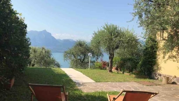 Eine Terrasse mit zwei Holzstühlen, die auf einen See blicken, umgeben von Grünpflanzen und blauem Himmel.