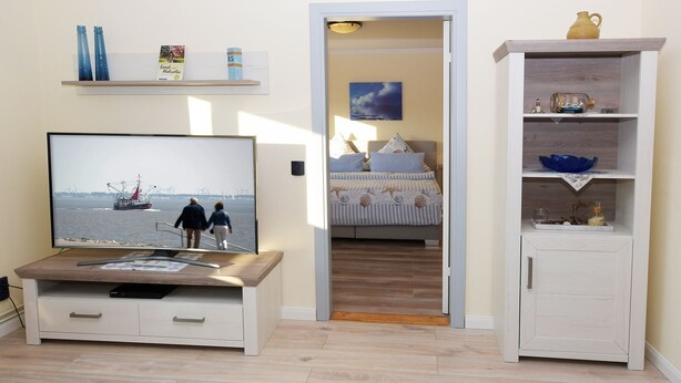 A room with a TV displaying a scene of two people walking by the sea, a bed with a striped bedspread, and a white cabinet with shelves. - Ferienwohnung Hummer (Büsum)