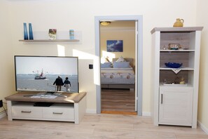 Una habitación con una televisión mostrando una escena de dos personas caminando por la playa, una cama con colcha a rayas y un armario blanco con estantes.