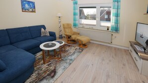A living room with a blue sofa, a yellow armchair, and a small table. There's a window with curtains, a radiator, and a TV on a wooden stand. - Ferienwohnung Hummer (Büsum)