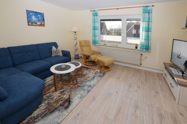 Una sala de estar con un sofá azul, un sillón amarillo y una mesita. Se aprecia una ventana con cortinas, un radiador y un televisor sobre un mueble de madera.