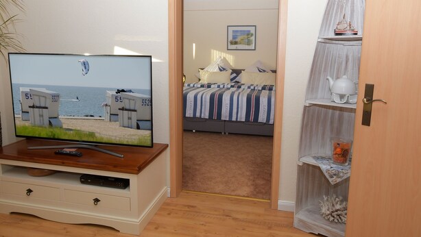 Een slaapkamer met een televisie op een houten standaard, een bed met streepjesbeddengoed, een deur naar een andere kamer en een plank met decoratieve objecten.