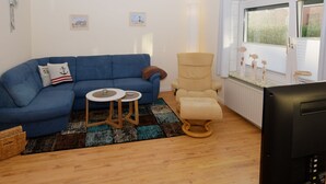 Een woonkamer met een blauwe bank, een beige fauteuil, twee kleine ronde tafels en een televisie.