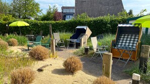Een tuin met buitenmeubilair, waaronder stoelen en een tafel, omringd door zand en beplanting.