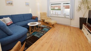 Een woonkamer met een blauwe bank, een beige fauteuil, een klein rond tafeltje en een raam met gordijnen.