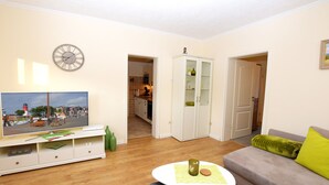 Ein Wohnzimmer mit Fernseher, Sofa, Couchtisch und Esstisch. Durch eine offene Tür ist eine Küche zu sehen.