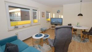 A living room with a sofa, a television, a dining table with chairs, and large windows with yellow blinds.