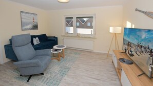 A living room with a grey armchair, a blue sofa, a small round table, a floor lamp, and a painting on the wall. - Vacation apartment Robbe in house Alina (Büsum)