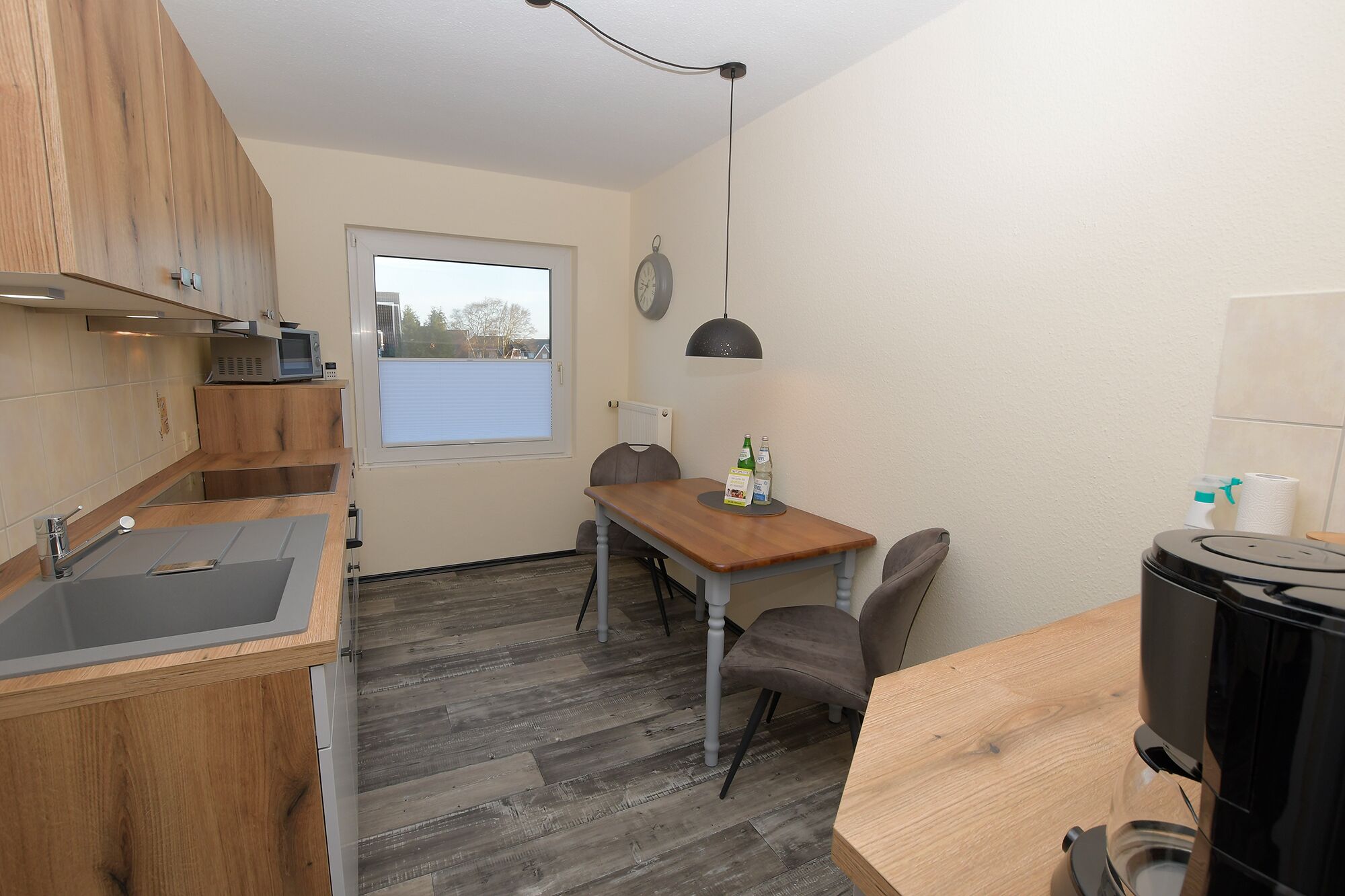 A compact kitchen with wooden cabinets, a built-in microwave, a dining table with chairs, a window with blinds, and a clock on the wall.