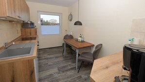 A compact kitchen with wooden cabinets, a built-in microwave, a dining table with chairs, a window with blinds, and a clock on the wall. - Vacation apartment Robbe in house Alina (Büsum)
