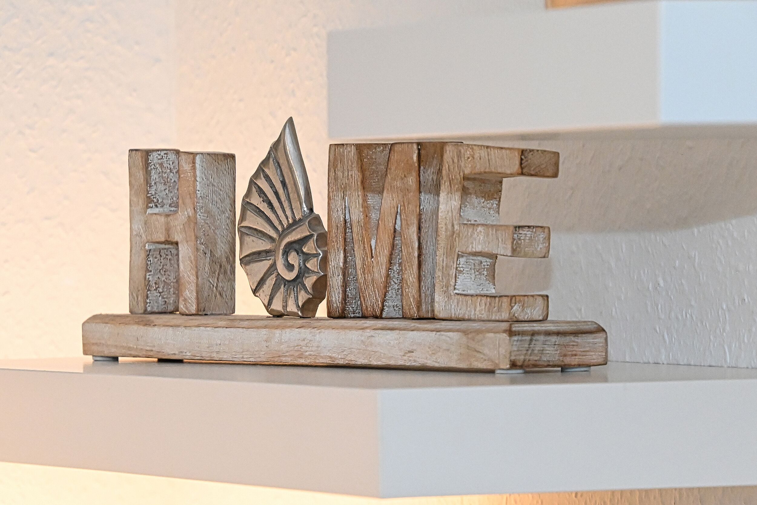A wooden word art piece spelling "HOME" with a bird design, placed on a white shelf against a textured wall.
