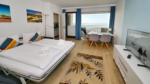 A hotel room with a bed, a desk, and a TV. There is a view of the beach through the window. - Herzlich Willkommen in der der Ferienwohnung "meerblick 9"! (Büsum)