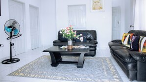A modern living room with a black leather sofa, a black coffee table, and a standing fan. - Luxury 2 Bed Self Catering Apartment in Masvingo (Masvingo)