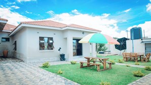 A white building with a tiled roof, a paved patio, and a small lawn with picnic tables and chairs. - Luxury 2 Bed Self Catering Apartment in Masvingo (Masvingo)