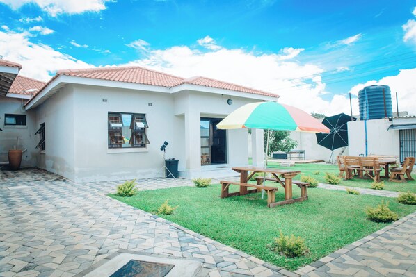 A white building with a tiled roof, a paved patio, and a small lawn with picnic tables and chairs. - Luxury 2 Bed Self Catering Apartment in Masvingo (Masvingo)