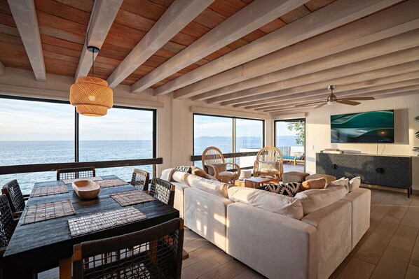 Un vaste espace de vie avec une grande fenêtre donnant sur la mer, une table à manger avec des sets de table, un canapé et un téléviseur posé sur un meuble.