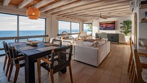 Un espai de vida ampli amb sostre de fusta, grans finestres amb vista al mar, una taula de dinar amb cadires i un sofà.