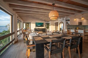 Un vaste espace de salle à manger avec une grande table et des chaises, une cuisine équipée d’appareils modernes et une vue sur la mer.
