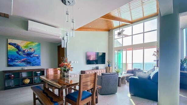 A modern living room with a wooden dining table, blue cushioned chairs, a large TV, and a view of the ocean through large windows.