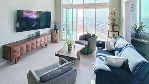 A modern living room with a large TV, comfortable seating, and a view through large windows.