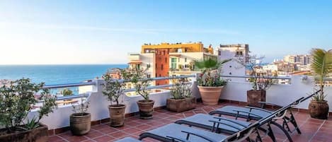 A rooftop terrace with lounge chairs, potted plants, and a view of the sea and buildings.