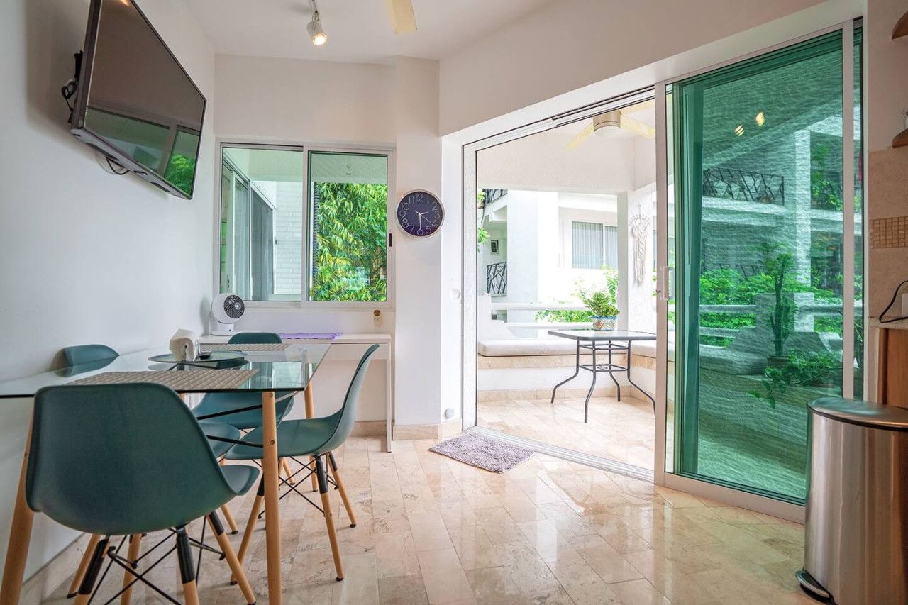 Et moderne køkken med indbygget ovn, mikroovn og kaffemaskine. Der er et spiseområde med et glasbord og stole, samt en stor skydedør i glas, der fører ud til en terrasse med udendørs møbler.