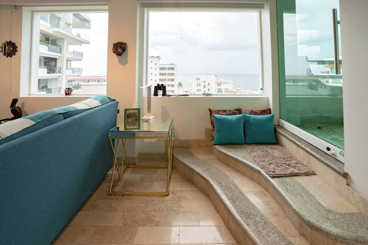 A living room with a blue sofa, a glass coffee table, a small table with a picture frame, a window with a view of buildings and the sea, and a set of stairs leading to a balcony.