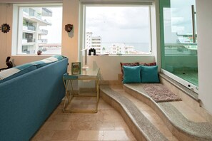 A living room with a blue sofa, a glass coffee table, a small table with a picture frame, a window with a view of buildings and the sea, and a set of stairs leading to a balcony.