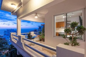 A balcony with a view of the sea and buildings, a ceiling fan, and a small table with a plant.