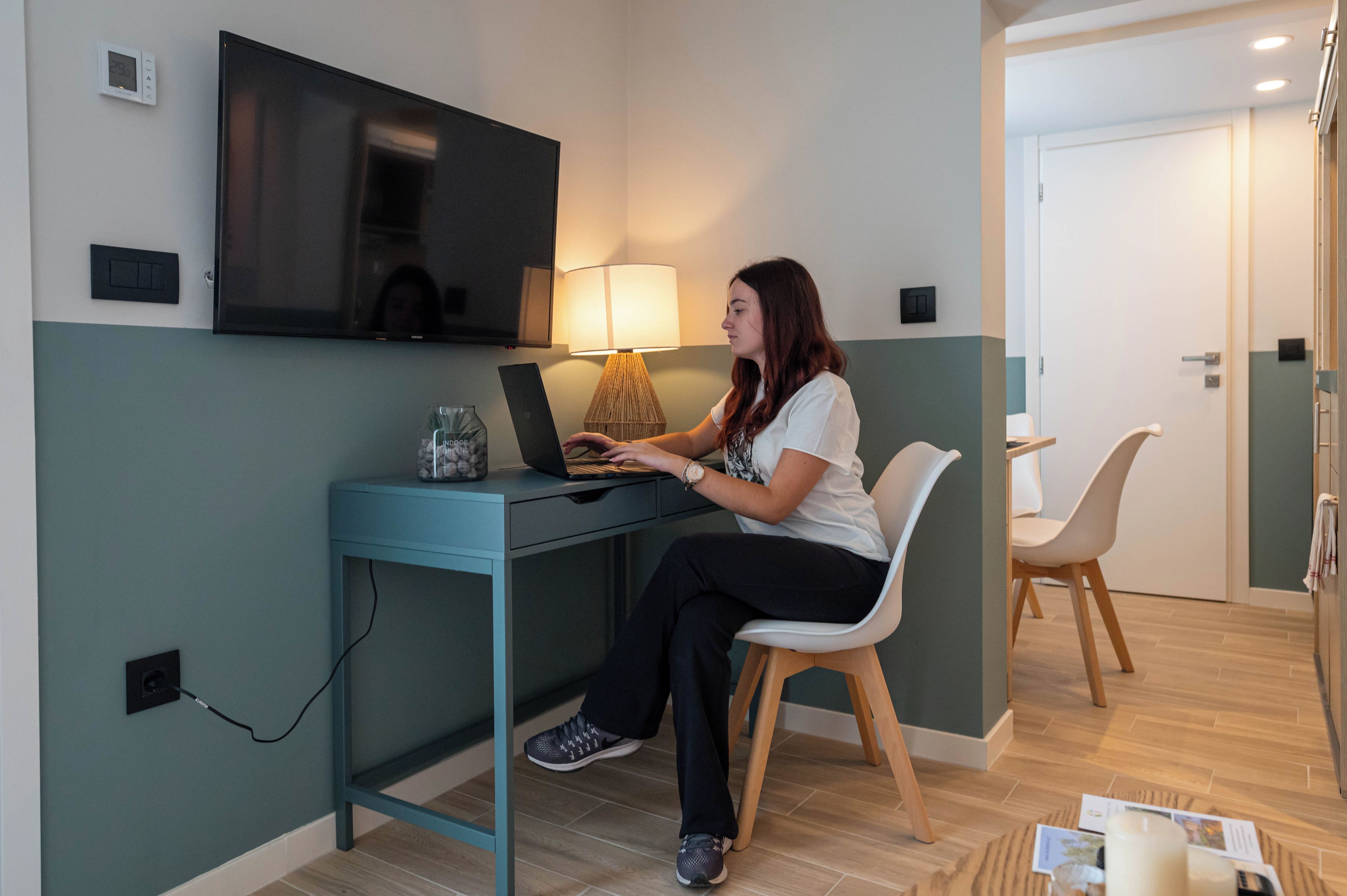 Una mujer sentada en un escritorio con una computadora portátil, un televisor montado en la pared y una lámpara sobre el escritorio.