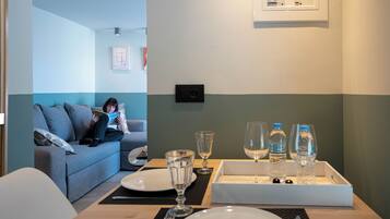 A room with a dining table set for two, a sofa, and a wall-mounted light fixture.