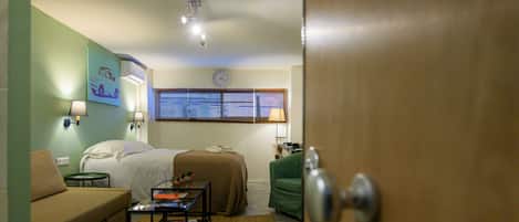 A room with a bed, a green armchair, a small table with books, a lamp, and a clock on the wall.