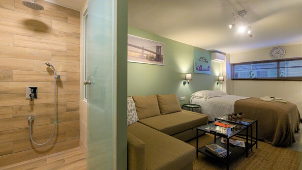 A hotel room with a shower, a sofa, a bed, a small table with items, and a wall clock. - Pols Studio - Project Kalamata Mediterranean Suites (Kalamata)