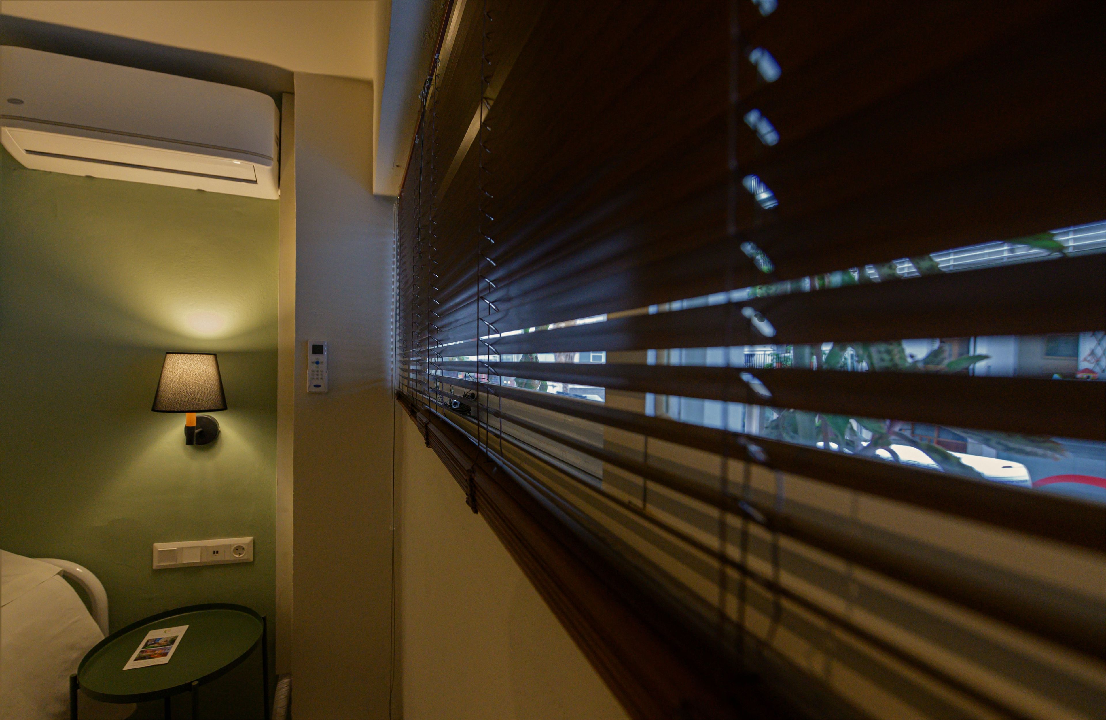 Una habitación de hotel con una cama, una mesita de noche con lámpara, una pequeña mesa con una tarjeta y una ventana con persianas.