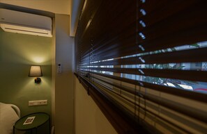A hotel room with a bed, a nightstand with a lamp, a small table with a card, and a window with blinds.