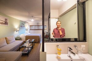 A hotel room with a bed, a sofa, a coffee table, a mirror, and a bathroom with a sink and mirror.