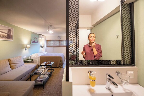 A hotel room with a bed, a sofa, a coffee table, a mirror, and a bathroom with a sink and mirror.