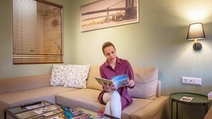 A person sitting on a sofa with a magazine, a glass coffee table with magazines and a duck decoration, a lamp, and a framed picture on the wall. - Pols Studio - Project Kalamata Mediterranean Suites (Kalamata)