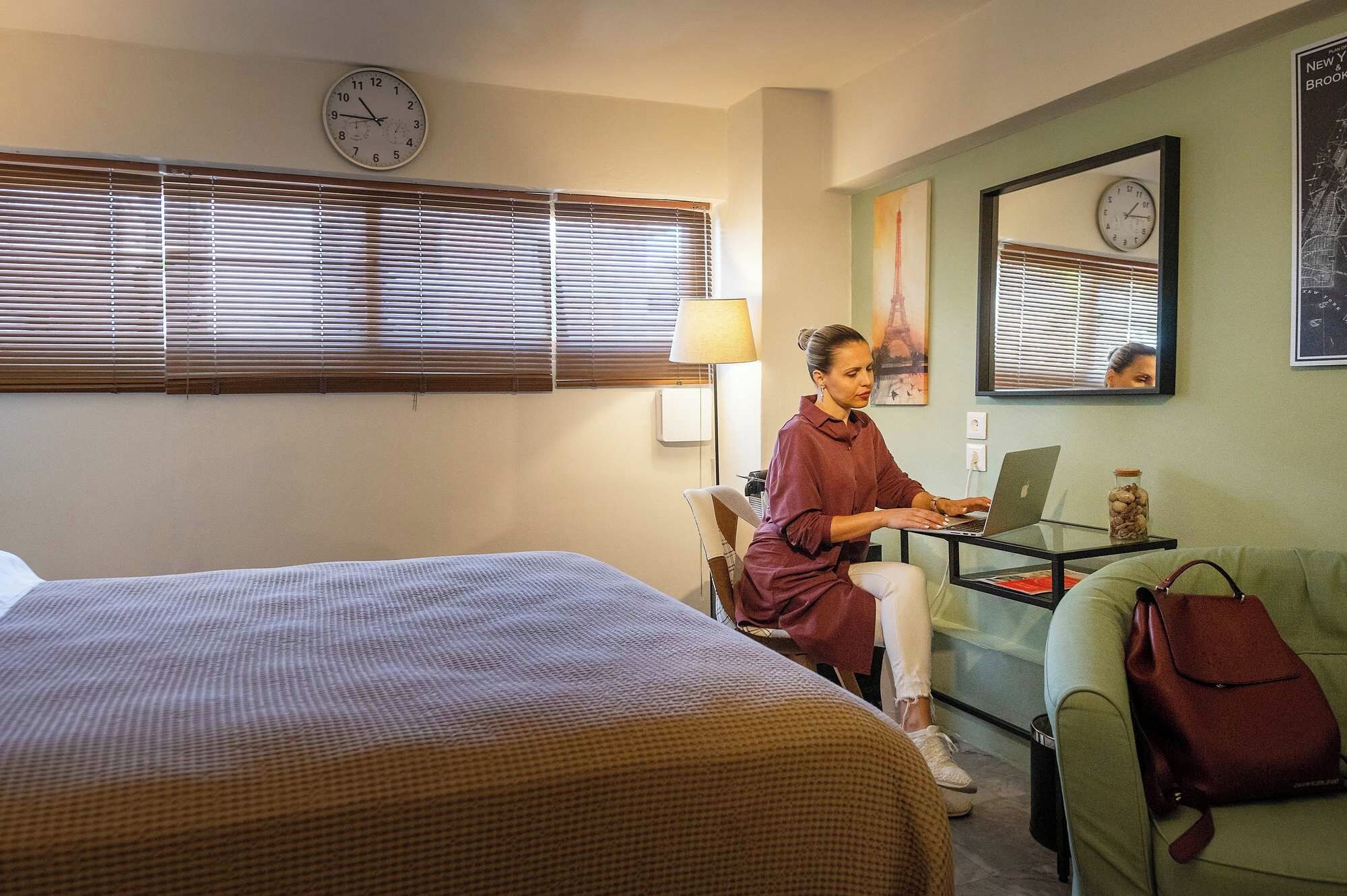 Una habitació d'hotel amb un llit, una taula de treball amb un ordinador portàtil, una cadira, un sofà i dos rellotges penjats a la paret.