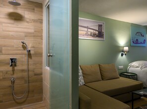 A hotel room with a shower, a bed, a sofa, a nightstand, and a wall-mounted lamp. - Pols Studio - Project Kalamata Mediterranean Suites (Kalamata)