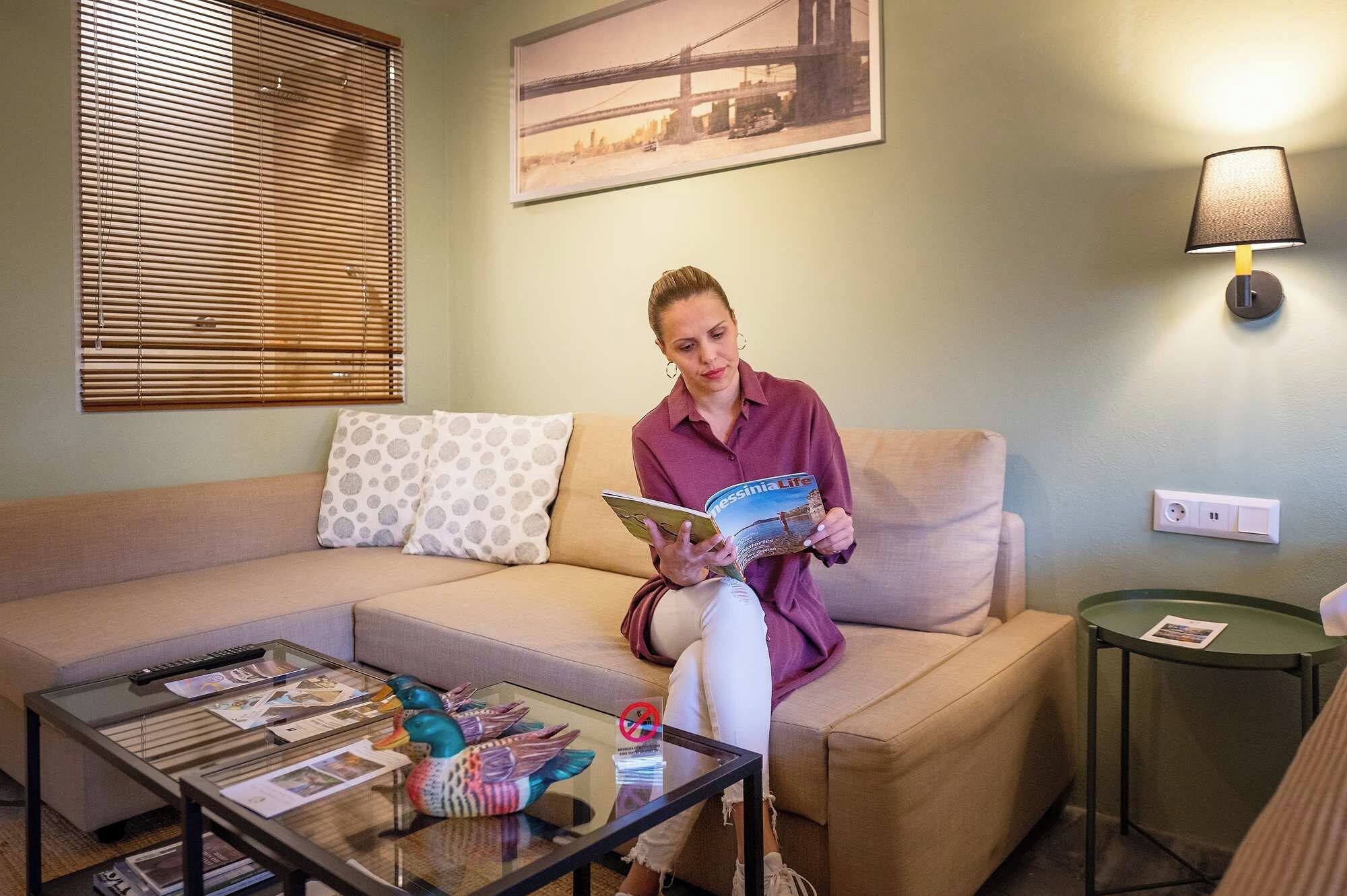 A person sitting on a sofa with a magazine, a glass coffee table with magazines and a duck decoration, a lamp, and a framed picture on the wall.