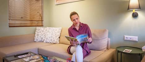 Una persona sentada en un sofá con una revista, una mesita de centro con revistas y una decoración de pato, una lámpara y un cuadro en la pared.